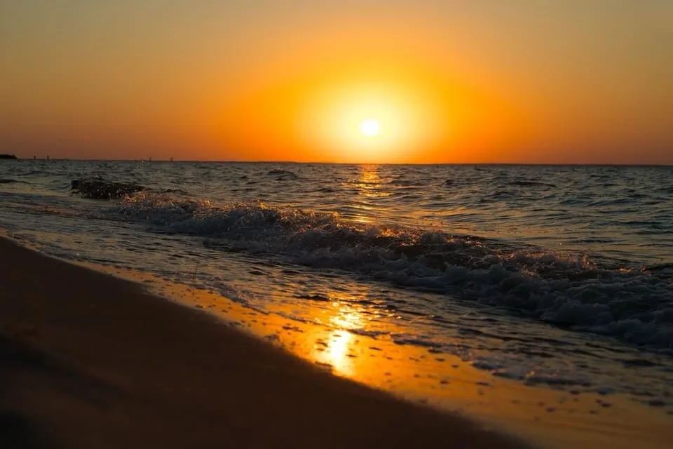 A serene sunset over the ocean with waves gently rolling onto a sandy beach, the sun casting a warm golden glow across the…
