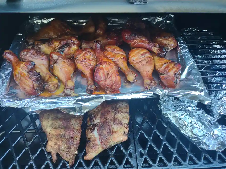 A close-up photo shows a grill with two levels of food. The top level holds a foil-lined tray full of glazed, cooked chicken…