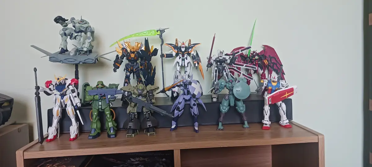 A wooden shelf displays a collection of various Gundam model kits, each painted and posed differently, standing on a black…