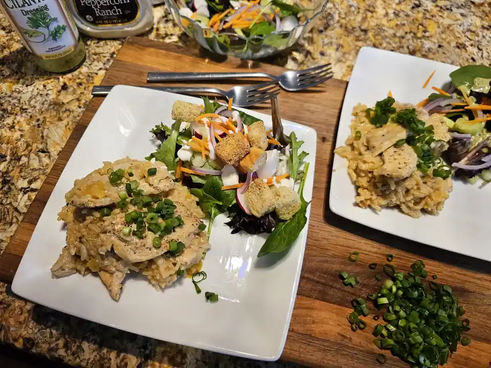 Two plated meals of chicken over rice with salad sides sit on a wooden cutting board. One plate has croutons and dressing…