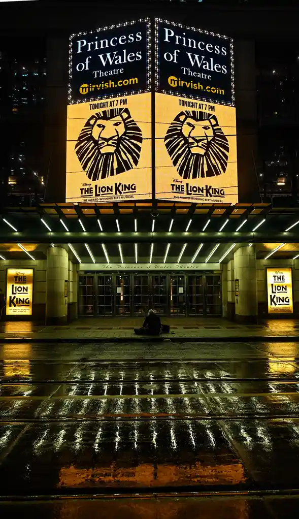 A person sits alone on the wet sidewalk in front of the brightly lit Princess of Wales Theatre at night, where large…
