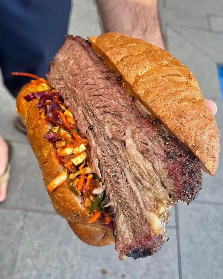 A person holds a large sandwich with a thick, juicy, sliced roast beef filling and a colorful coleslaw mix of shredded…
