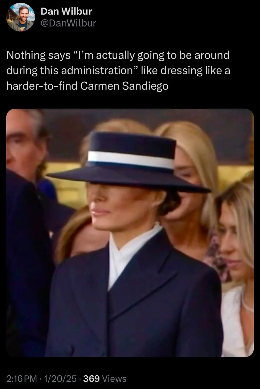 A person wearing a dark blue fedora with a white band and a matching dark blue suit, obscuring their eyes, stands in a…