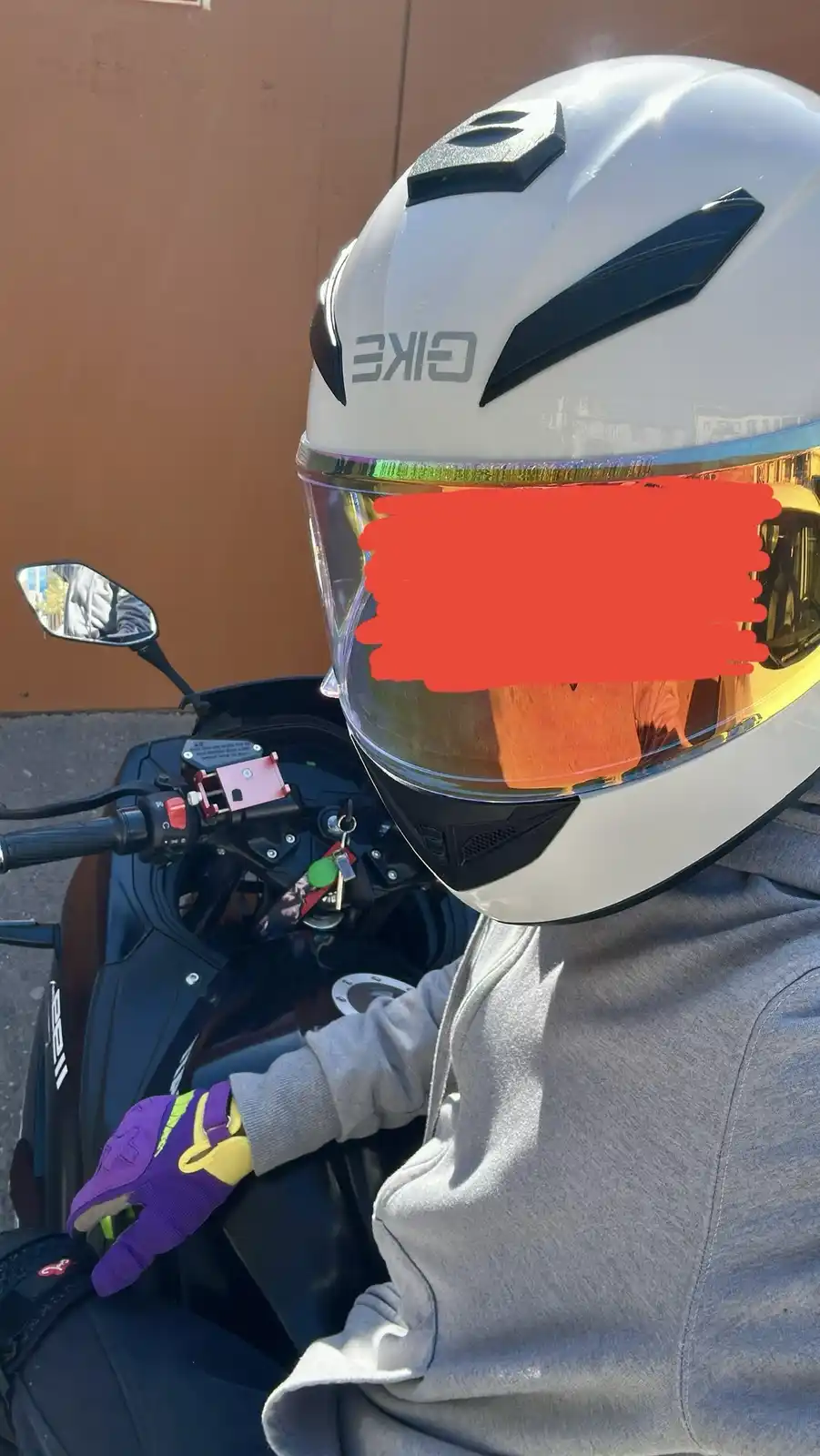 A person wearing a white GKE motorcycle helmet with a red-painted visor sits on a black scooter, wearing a grey hoodie and…