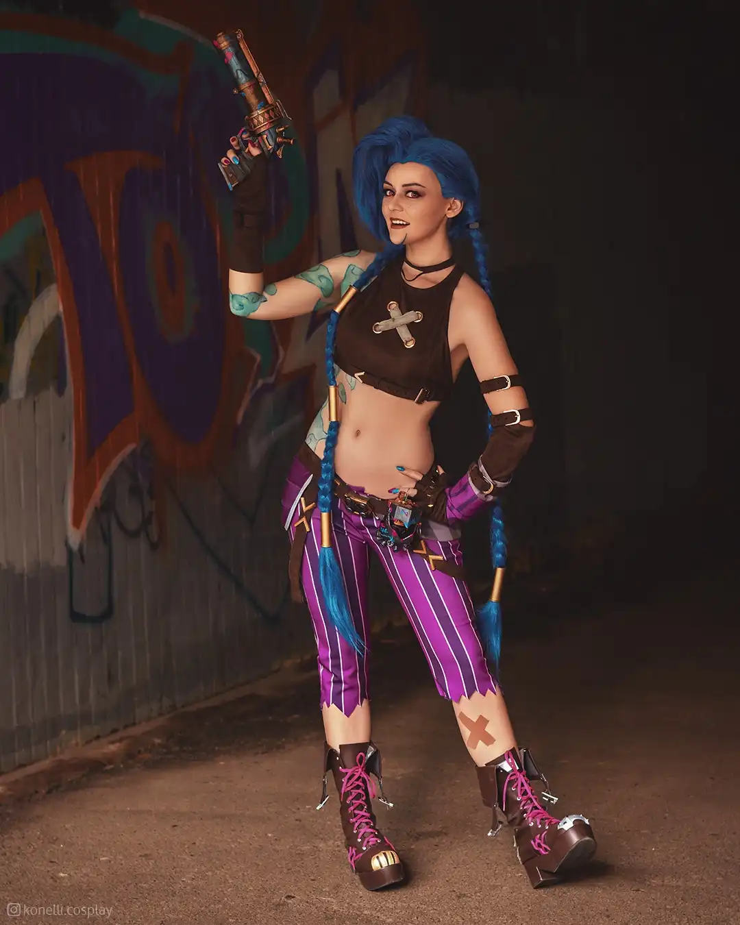 A cosplayer dressed as the video game character Jinx, holding a large ornate gun, poses confidently in front of a…