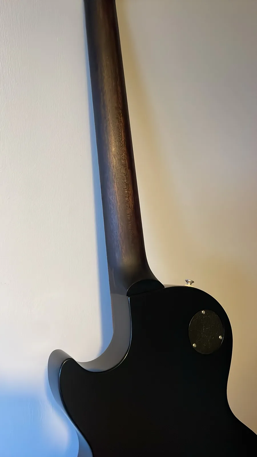 A close-up photo shows the back of a black electric guitar, focusing on the neck heel and body, leaning against a white wall…