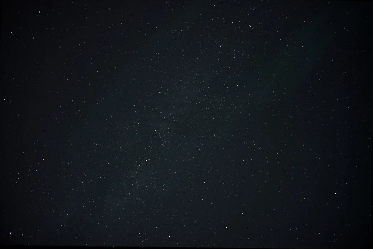 A dark night sky filled with countless stars and the faint, diffuse glow of the Milky Way galaxy stretching across the…
