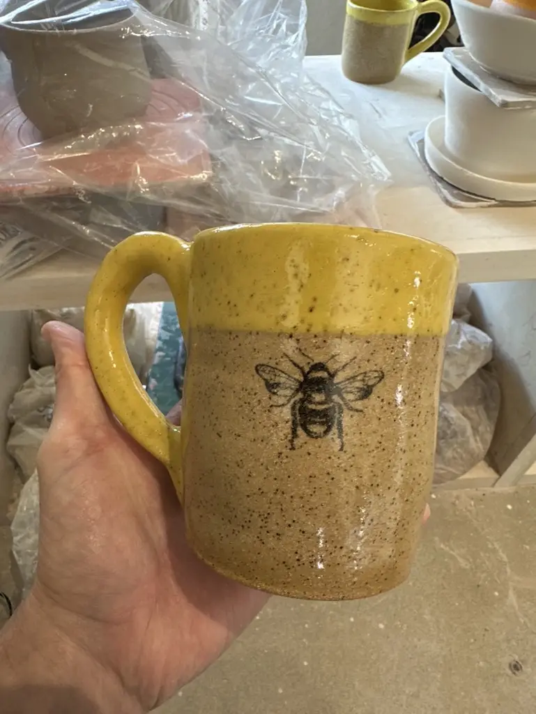 A hand holds a handmade ceramic mug with a yellow rim and speckled tan body, featuring a black bee illustration. The…