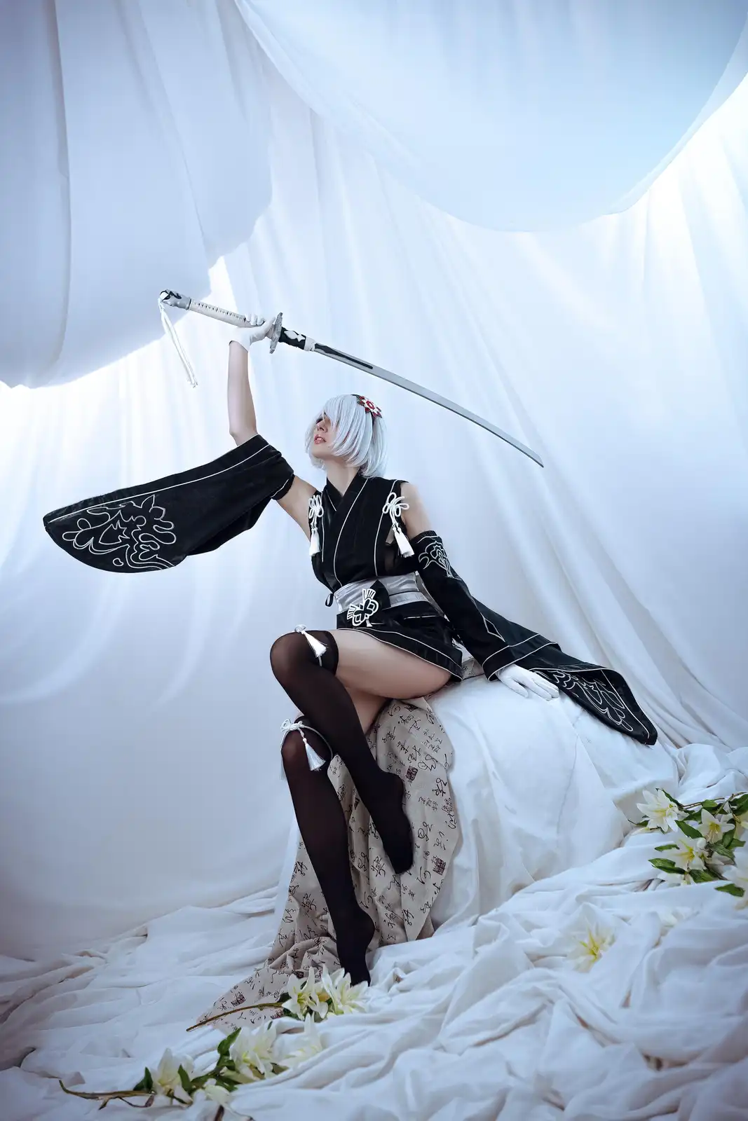A person in a black and white cosplay outfit with white hair, holding a katana aloft while seated on a white draped surface…
