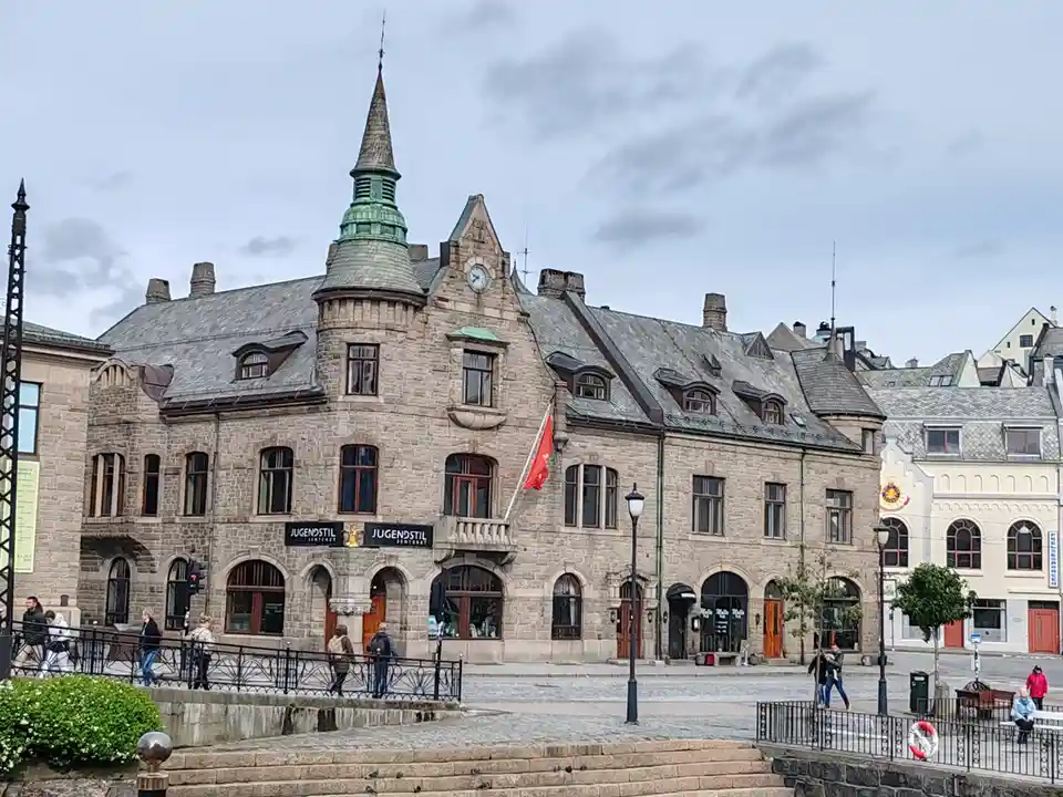 A wide shot of a large, ornate stone building with a clock tower and a red flag, labeled 'JUGENDSTIL SENTRUM' on its facade…