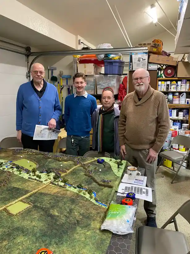 Four men stand behind a large table covered with a detailed miniature wargaming battlefield featuring tiny soldier figures…