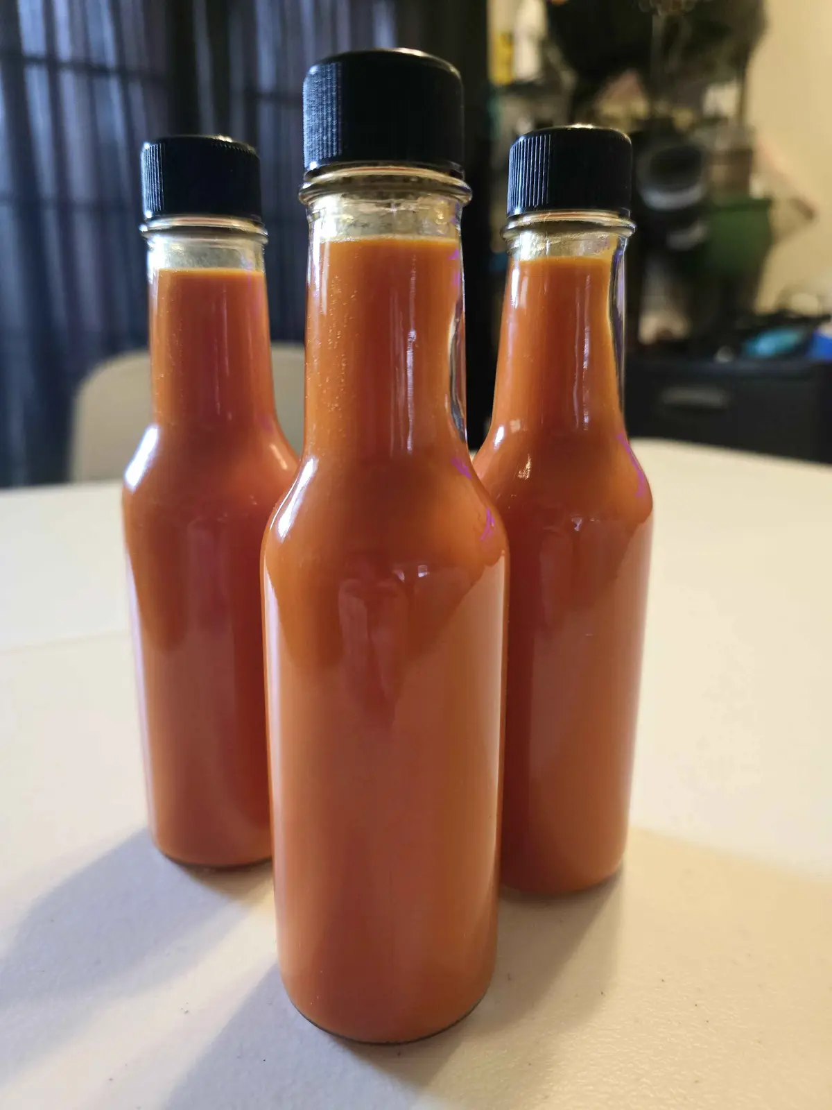 Three identical glass bottles filled with orange-red hot sauce, each capped with a black lid, are arranged in a row on a…