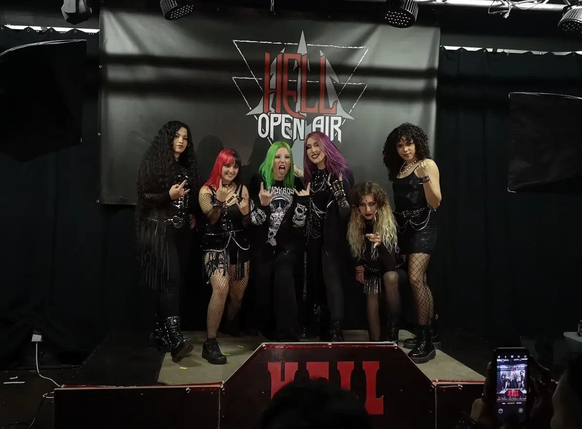 Six women in heavy metal concert attire, including black outfits, fishnet stockings, and brightly colored hair (red, green…