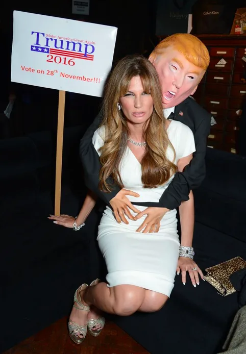 A woman in a white dress is kneeling on a couch while a person wearing a large Donald Trump mask hugs her from behind. A…