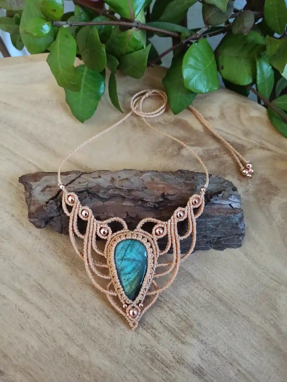 A handmade macrame necklace with a large, teardrop-shaped blue-green labradorite stone set in an ornate, peach-colored cord…