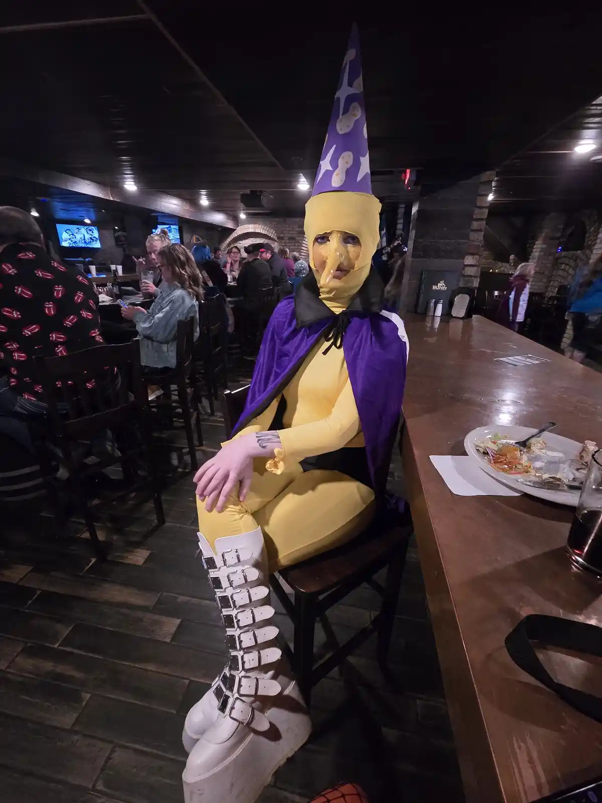 A person in a full-body yellow costume with a long nose, purple wizard hat, and purple cape sits at a restaurant table…