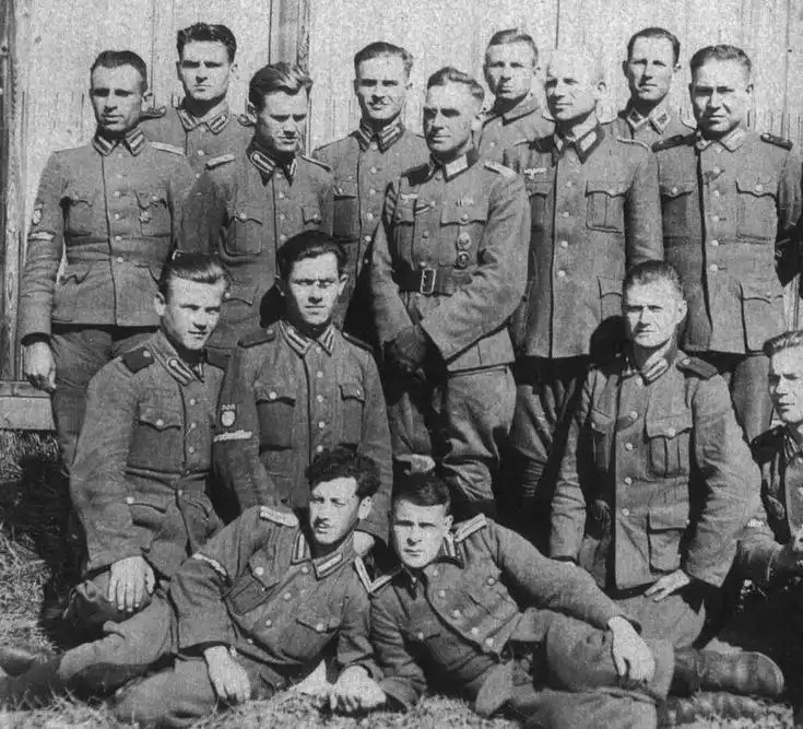 A black-and-white historical photo shows a group of men in military uniforms posing for a group photo, some standing and…