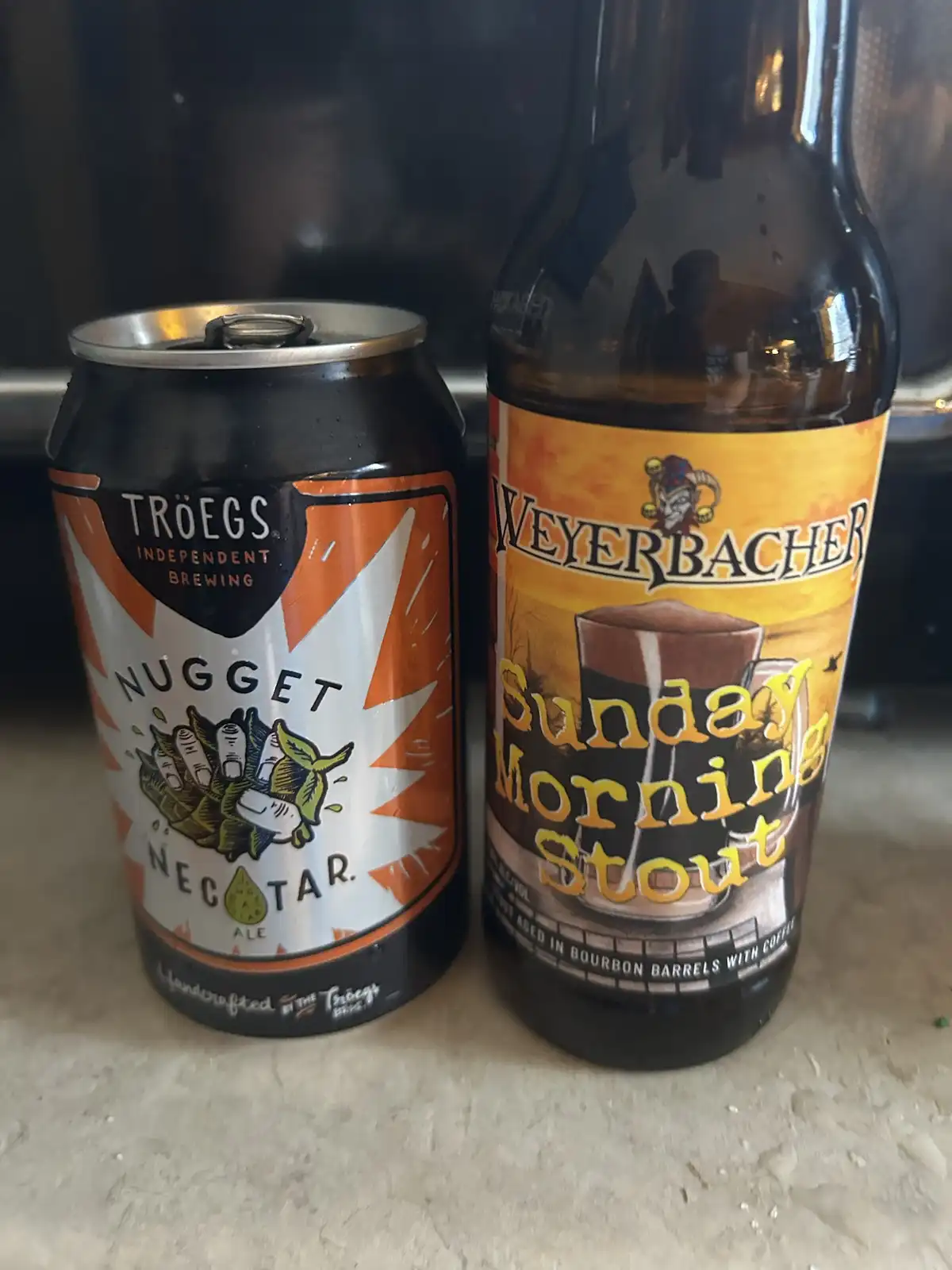 A black and orange can of Troegs Nugget Nectar Ale sits next to a brown bottle of Weyerbacher Sunday Morning Stout, both on…