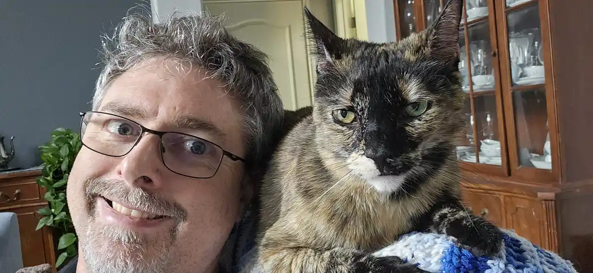 A man with glasses and a mustache smiles at the camera while holding a tortoiseshell cat that looks unimpressed. The cat…