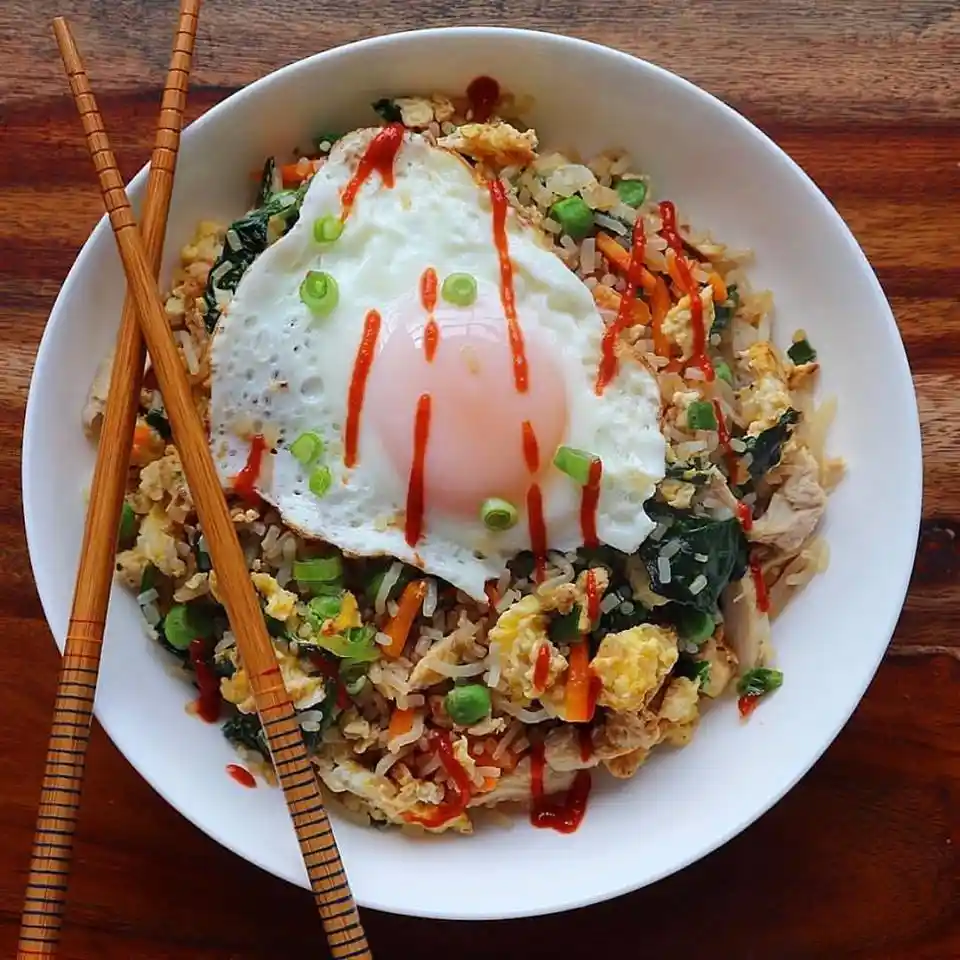 A top-down photo of a white bowl filled with fried rice mixed with vegetables, chicken, and a large sunny-side-up egg…