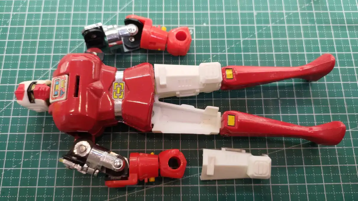 A red and white plastic robot toy is disassembled and laid out on a green cutting mat, with its limbs and torso separated…