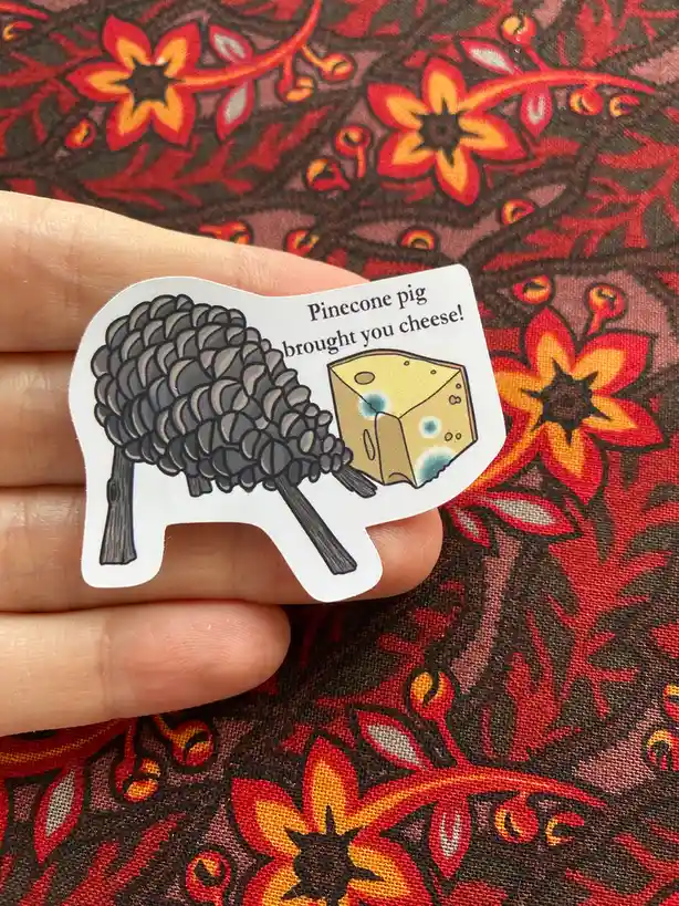 A hand holds a die-cut sticker depicting a whimsical creature shaped like a pig with a pinecone for a head, carrying a wedge…