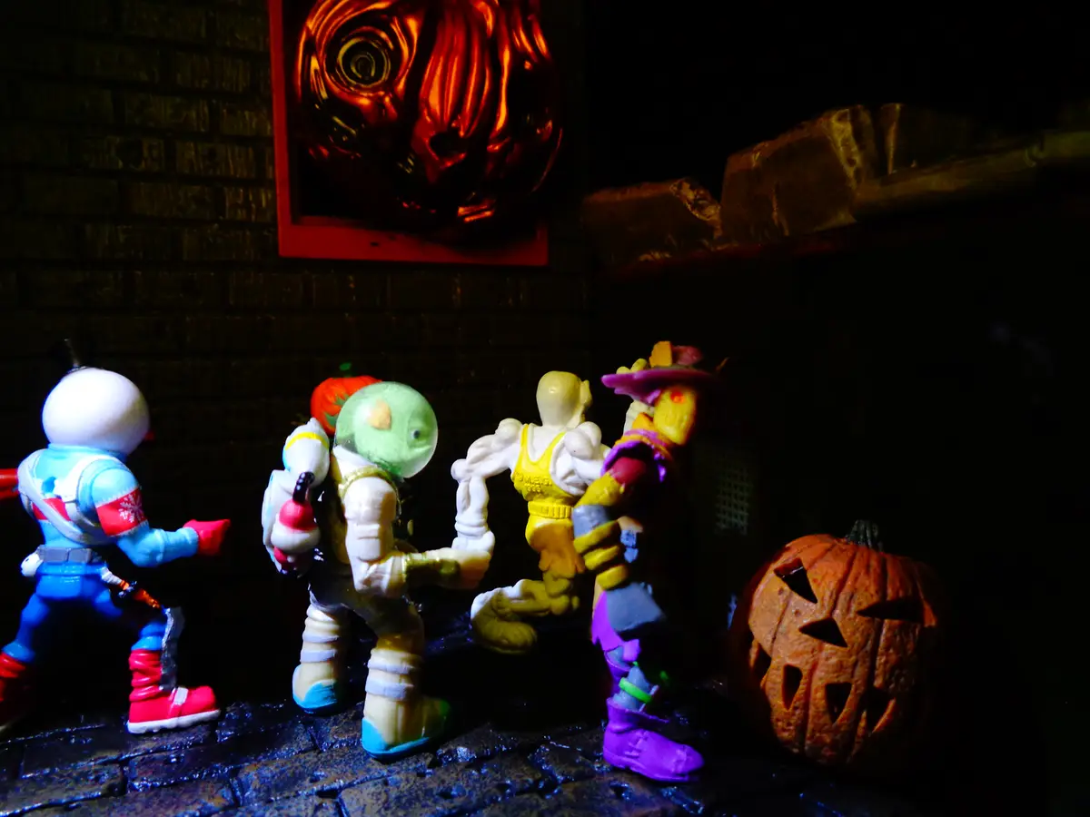 A dimly lit diorama shows four action figures standing on a cobblestone surface in front of a brick wall. A carved…
