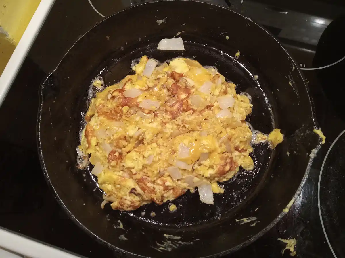 A close-up photo shows scrambled eggs mixed with chopped onions and melted cheese cooking in a black cast-iron skillet on a…