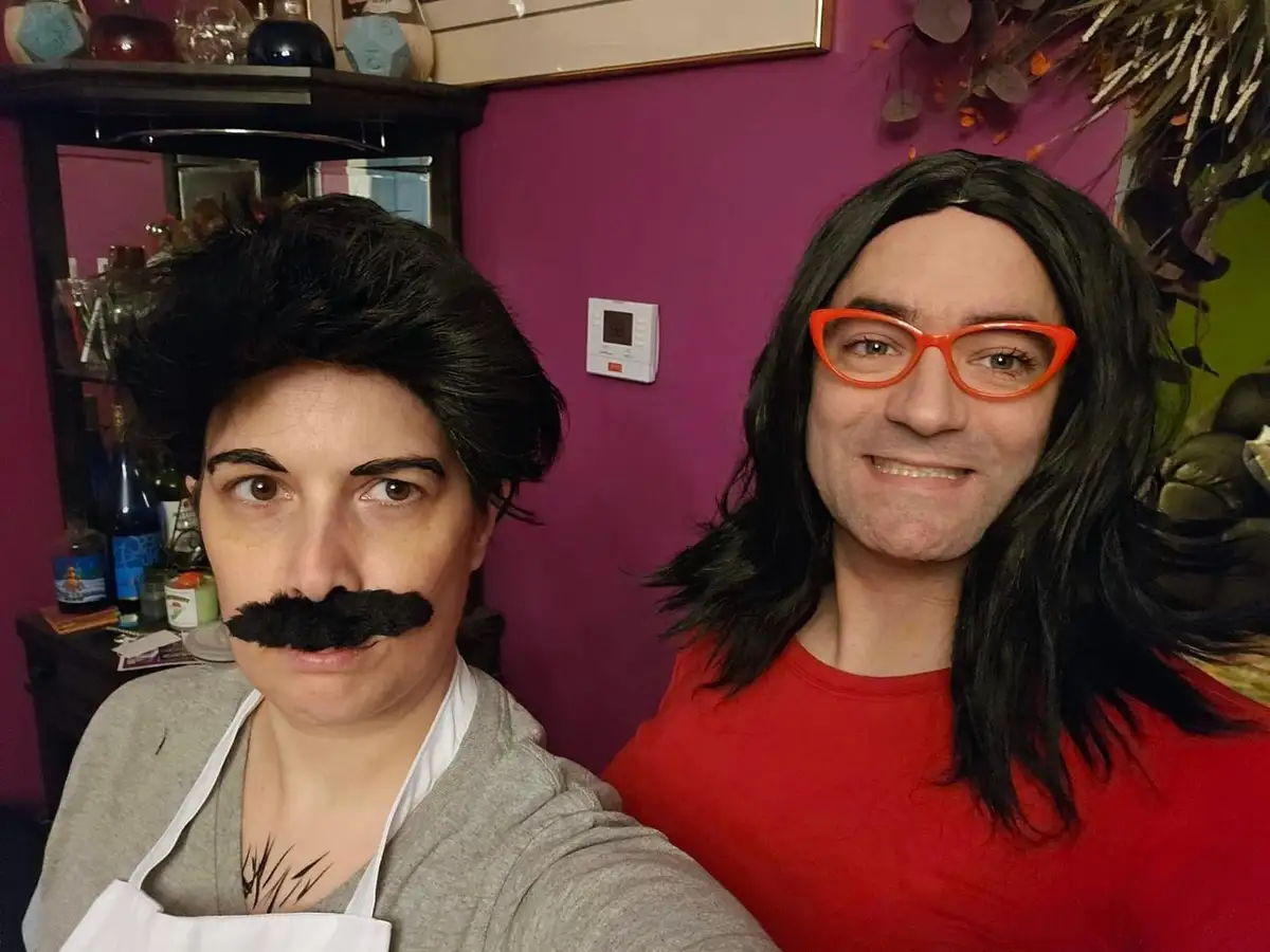 Two people pose for a selfie in a room with purple walls; the person on the left wears a grey sweater, white apron, and a…