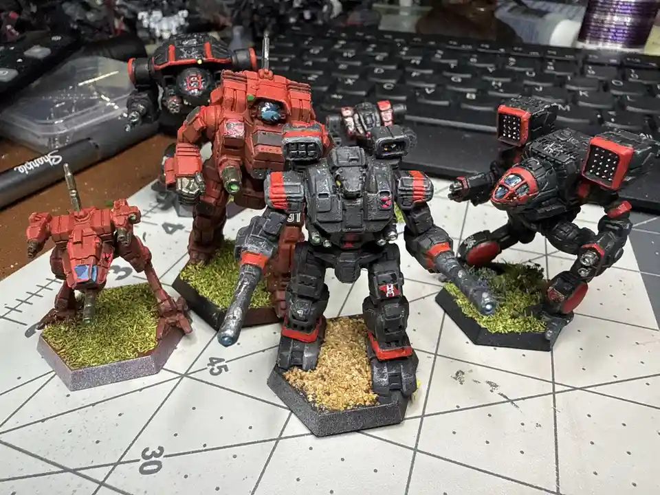 A photo shows five painted miniature mechs on hexagonal bases with grass or sand textures, arranged on a grid-lined gaming…