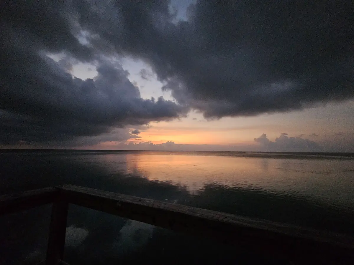 A dramatic sunset over a calm ocean, viewed from a wooden pier railing, with dark storm clouds overhead and orange light…