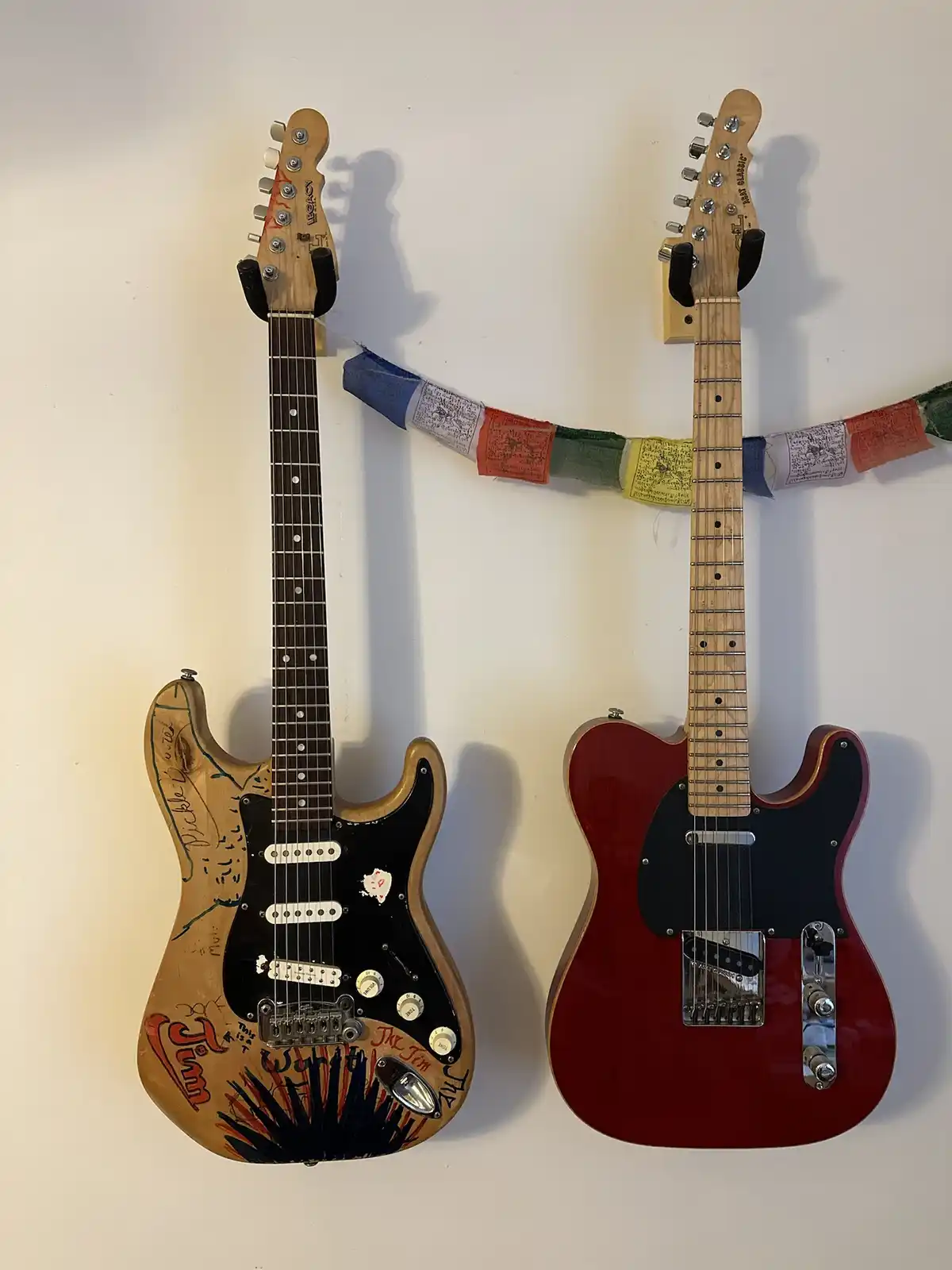 Two electric guitars hang side-by-side on a white wall, connected by a string of colorful prayer flags. The guitar on the…