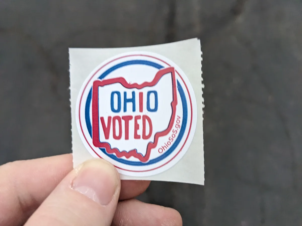 A close-up photo shows a hand holding a circular 'OHIO VOTED' sticker, which features a red outline of the state of Ohio…