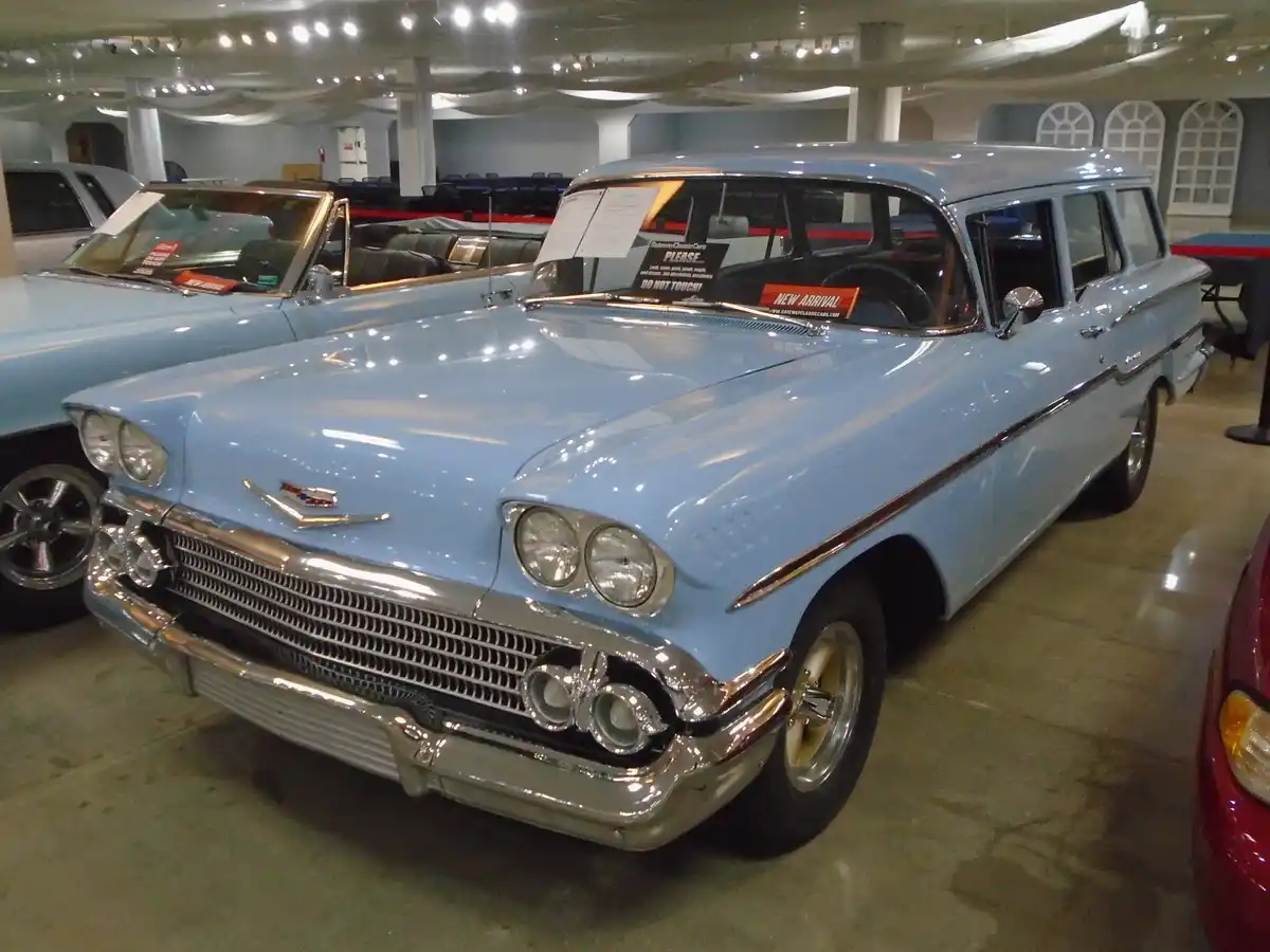 A light blue 1950s Chevrolet station wagon is displayed indoors in a car showroom, with a 'PLEASE... Look, admire…