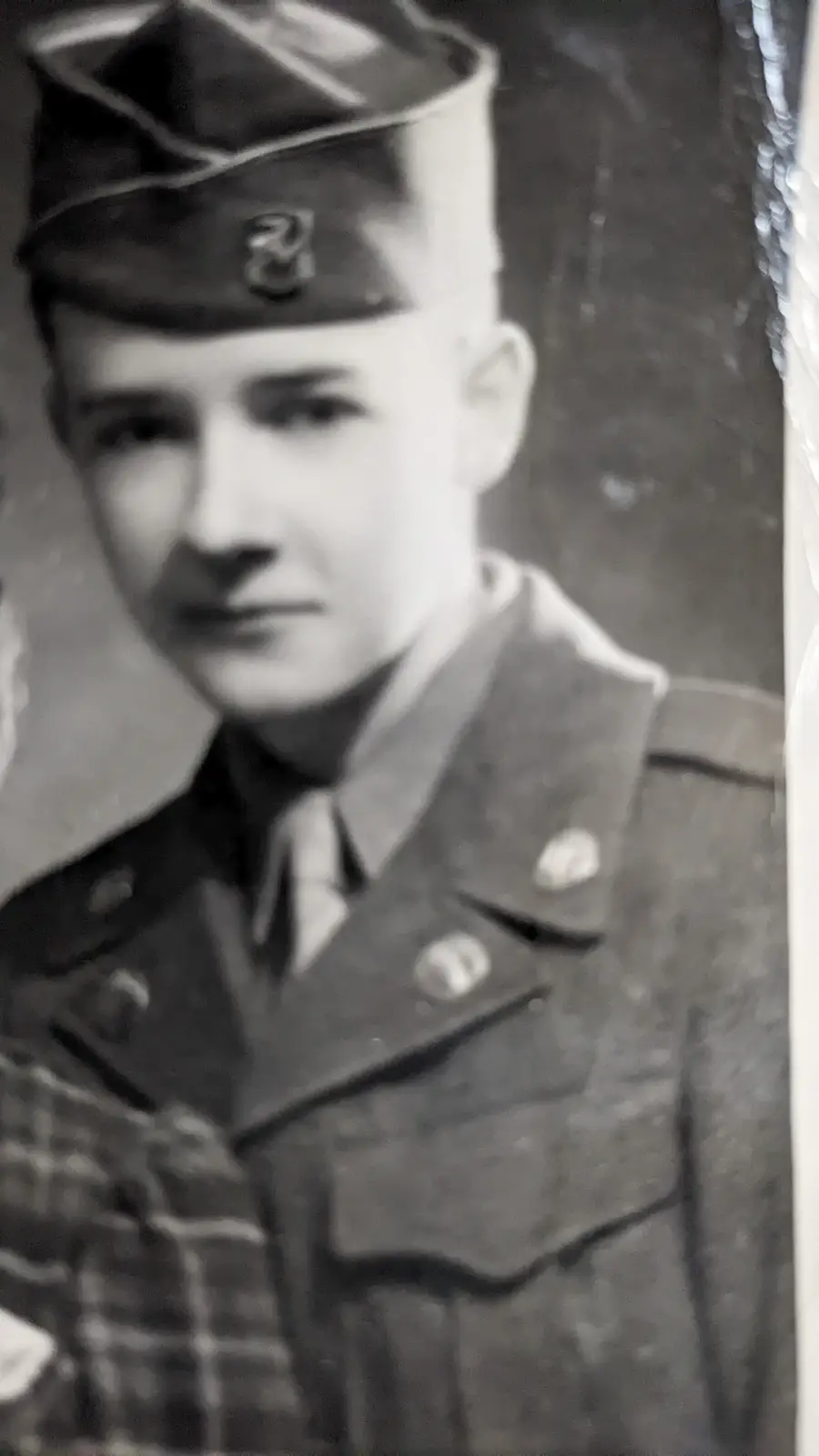 A grainy black-and-white portrait of a young man in a military uniform with a cap and insignia, looking directly at the…