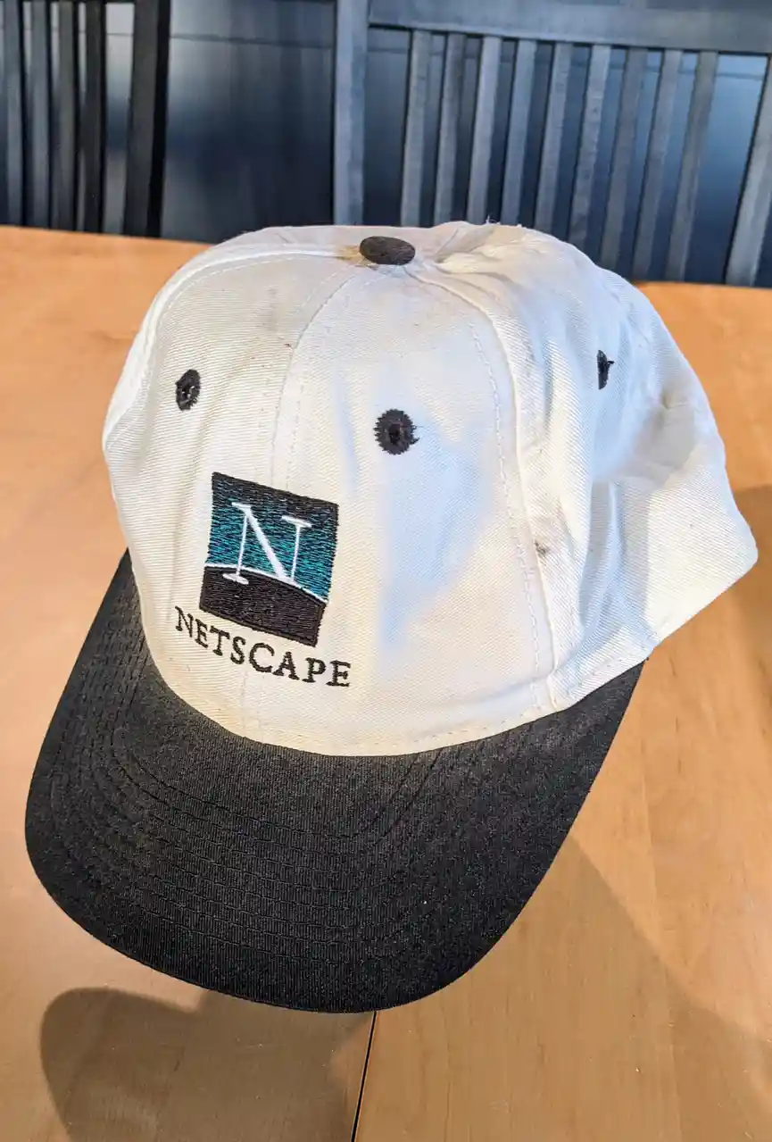 A white and black baseball cap with a faded Netscape logo and text sits on a wooden table, showing signs of wear and tear…