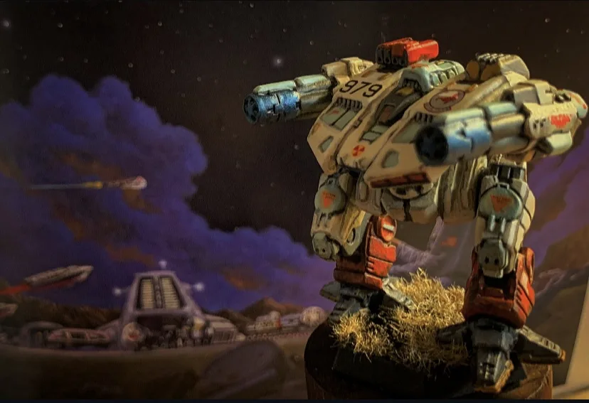 A painted miniature of a large, bipedal mech with the number 979 on its chest stands on a base with hay, set against a…