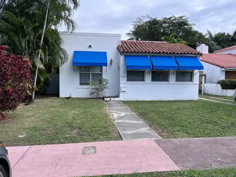 A white stucco house with a red-tiled roof and bright blue awnings over windows and a door, set in a grassy yard with palm…