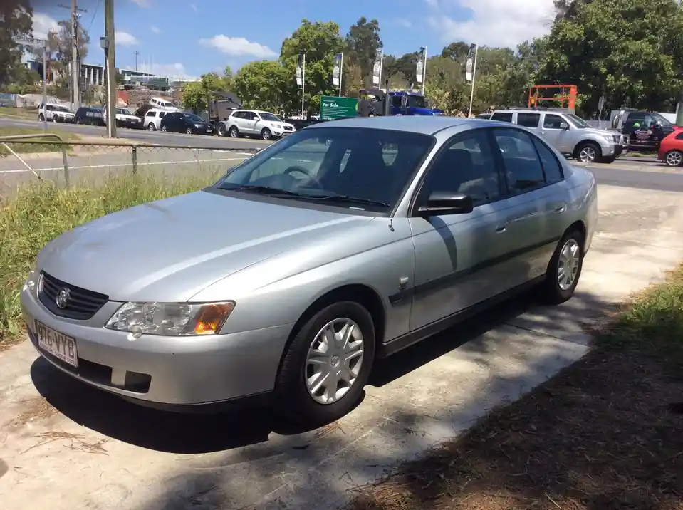 A silver Holden Commodore sedan is parked on a concrete surface next to grass, with other vehicles and a 'For Sale' sign…