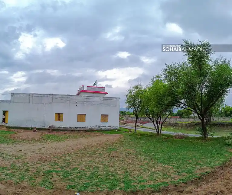a plain white single-story building with yellow windows and a red roofline, flying a green flag, sits in a rural setting…