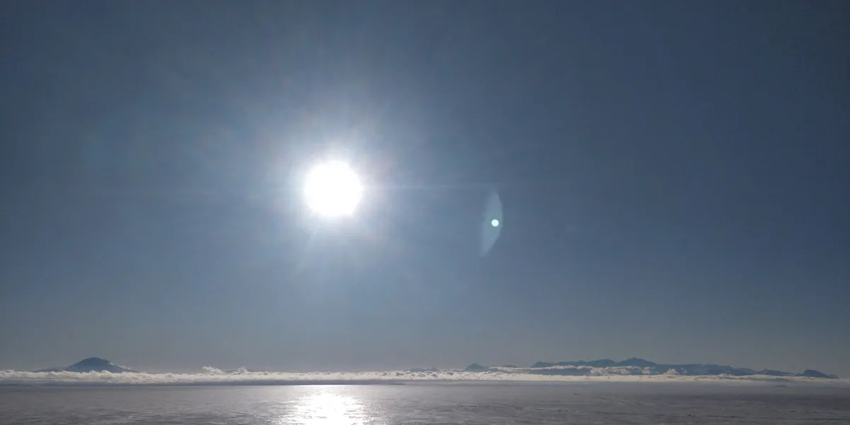 A bright sun shines over a vast, icy landscape with distant mountains on the horizon, partially obscured by a thin layer of…