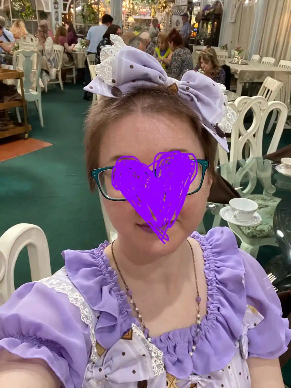 A person with short brown hair wears a large lavender bow headband and a matching ruffled lavender dress with lace trim…