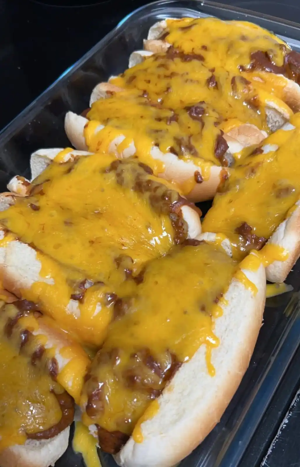 A close-up photo shows several chili cheese dogs in a glass baking dish, each topped with a thick layer of melted yellow…