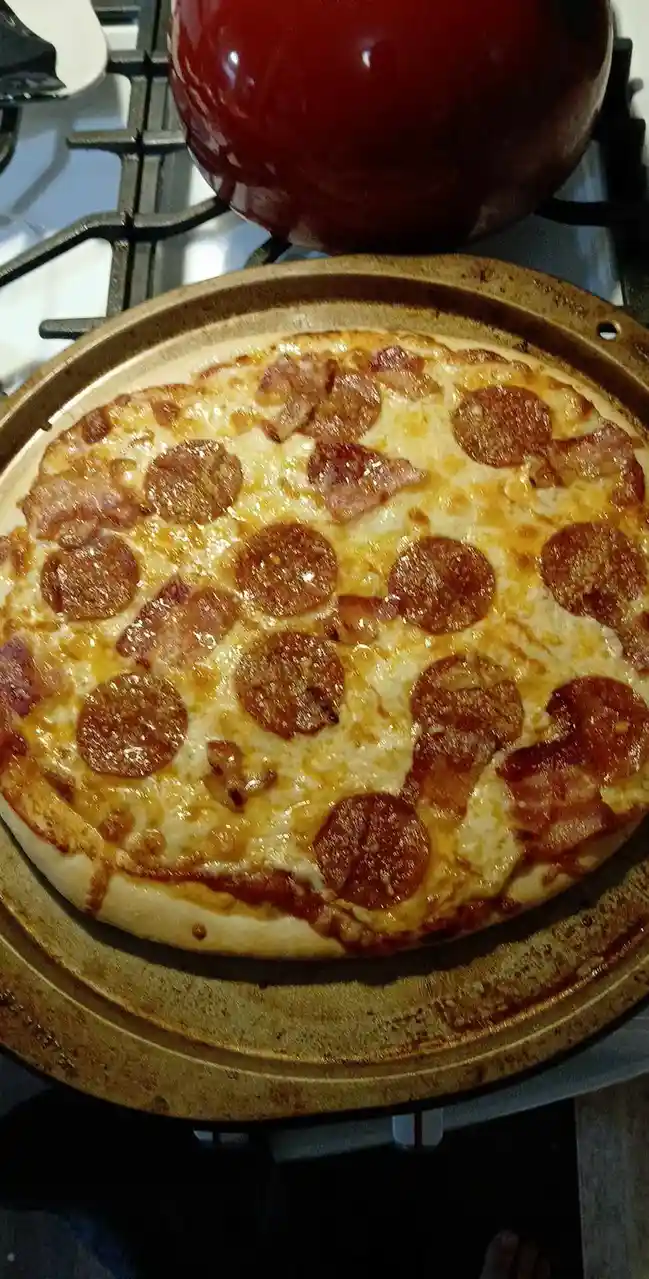 A freshly baked pepperoni and bacon pizza sits on a round, well-used baking stone on a stovetop, with a large red pot…