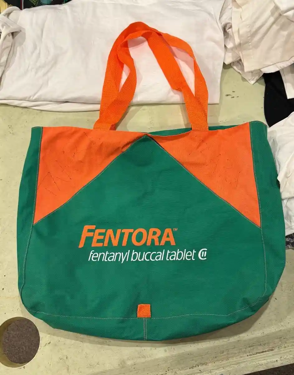 A green and orange tote bag with the text 'FENTORA fentanyl buccal tablet ™' printed on it, sitting on a surface with white…
