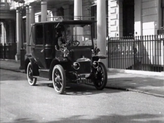 A black-and-white photo shows an old-fashioned, early 20th-century automobile parked on a street with classical architecture…