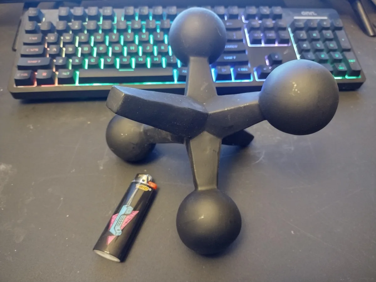 A black, four-sphere ergonomic desk toy sits on a dark desk next to a BIC lighter and in front of a backlit mechanical…