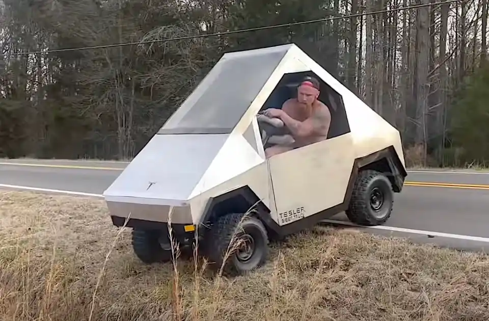 A shirtless, bearded man with a red headband drives a custom, angular, silver Tesla Cybertruck replica that is partially…