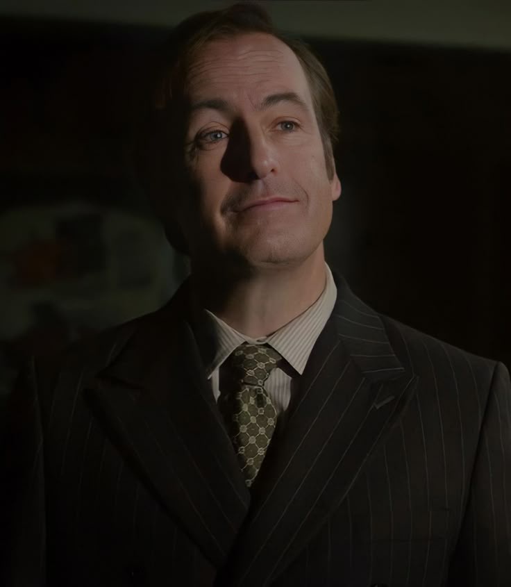 a man in a dark pinstripe suit and patterned tie looks upward with a subtle, knowing smirk under dim lighting. The overlaid…