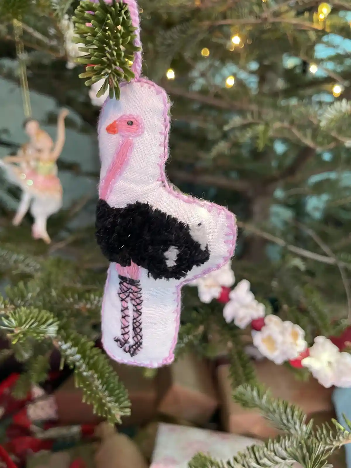 A handmade fabric ornament shaped like a pink and black bird, hanging on a Christmas tree with lights and other decorations…